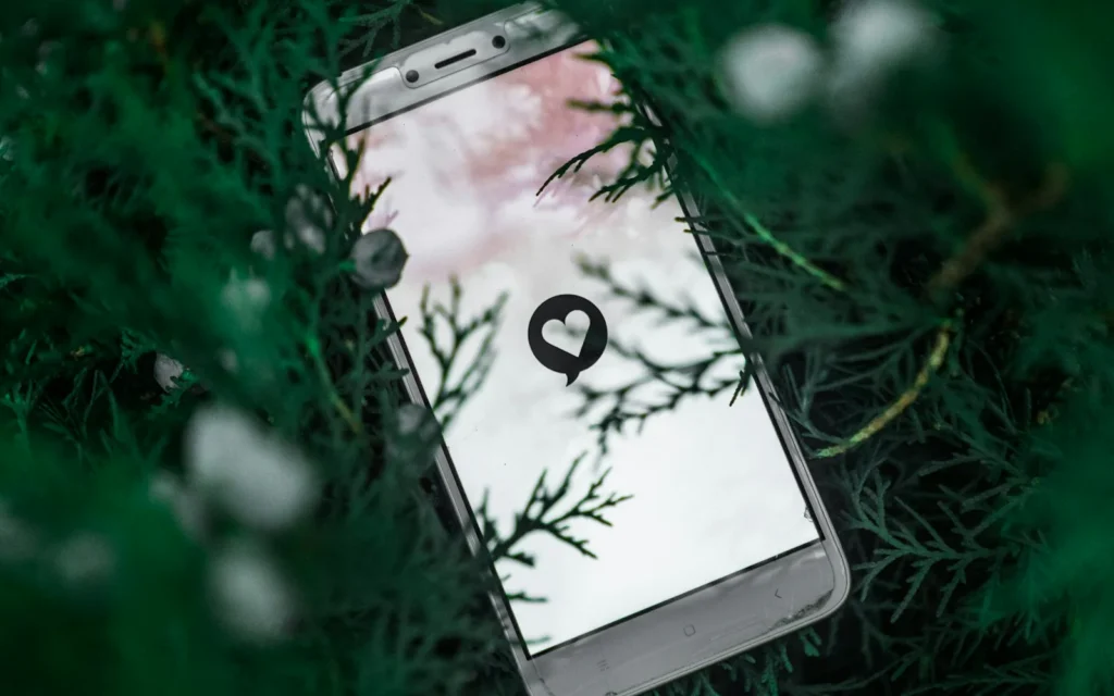A white smartphone with a heart-and-message icon lies among green foliage.