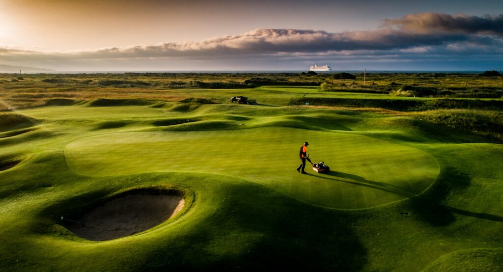 Greenkeeper mowing a golf putting green in low morning light.