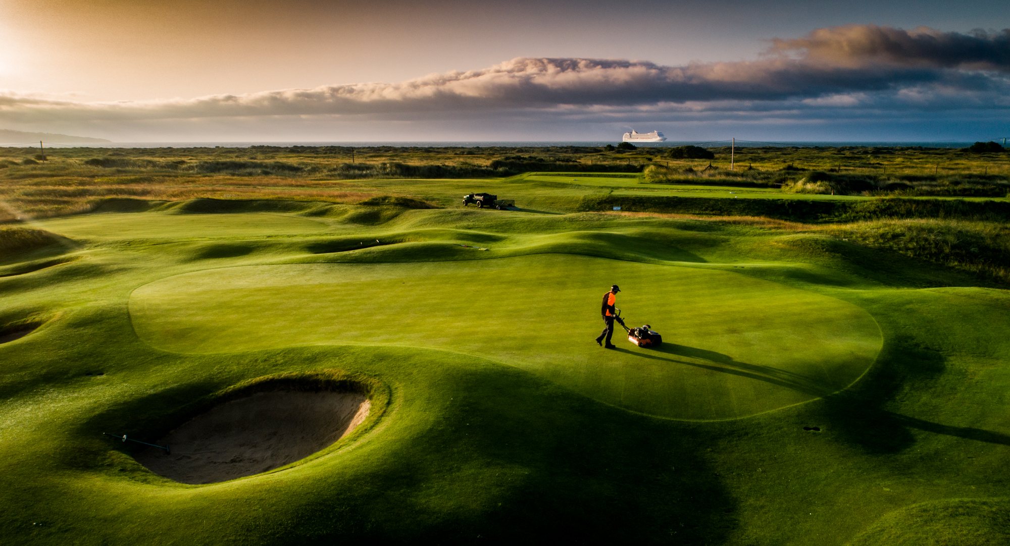 Greenkeeper mowing a golf putting green in low morning light.