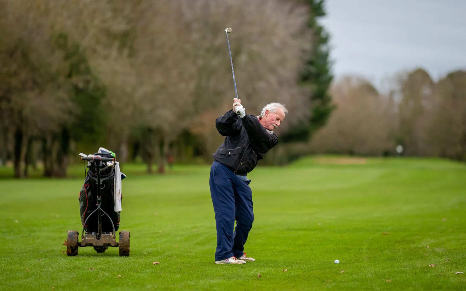 An older golfer swings beside a push trolley on a parkland course.